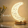 Crystal Table Lamp: The Crystal Table Lamp features a modern moon-shaped design atop a wooden base elegantly labeled “Moonlight.” This bedside lamp emits warm light, offers convenient touch controls for color changing and dimming, and pairs beautifully with yellow flowers in a glass vase placed nearby.
