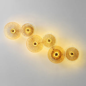 Six Amber Deco Wall Sconces, featuring circular gold wire designs, are mounted in a staggered line on a plain gray wall, casting a warm yellow glow.