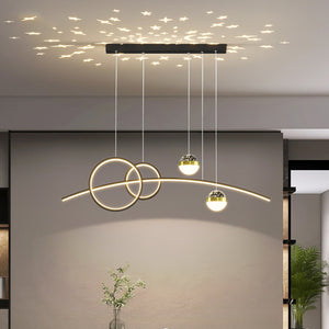 The Stellar Pendant Light features circular and linear elements that create star-shaped patterns with celestial accents when hung from the ceiling. A green plant can be seen in the background.