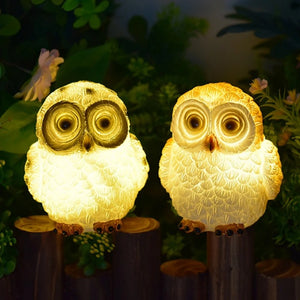 Two Solar Owl Lights rest on a wooden surface amid lush greenery and flowers, casting a warm glow in the darkness—a charming addition to any cottage garden décor.