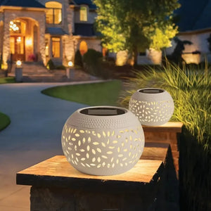 Two Solar Orb Lamps with leaf-pattern cutouts glow atop stone pillars, illuminating a curved walkway that leads to a well-lit house at night.