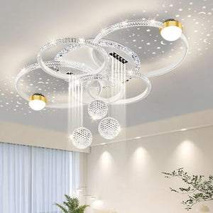 The Elegant Multi-Ring Ceiling Light features circular LED rings, hanging crystal spheres, and two gold-accented lights, casting star-shaped patterns on a white ceiling in a bright living room.