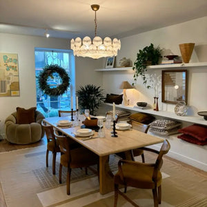A dining room with a light wood table set for six, modern chairs, an Italian Retro Pendant Light, wall shelves with decor, plants, and a window with a wreath.