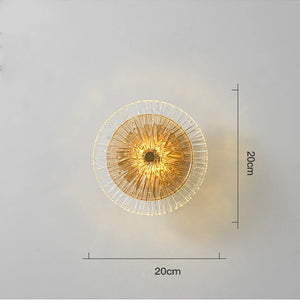 The Amber Deco Wall Sconce, with a gold center and clear glass disc, measures 20 x 20 cm and is stylishly displayed on a plain light-colored wall.