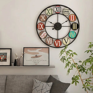 A Nordic Color-Ring Wall Clock with mixed numeral styles hangs above a gray couch, next to framed art and a potted plant on the nearby shelf.