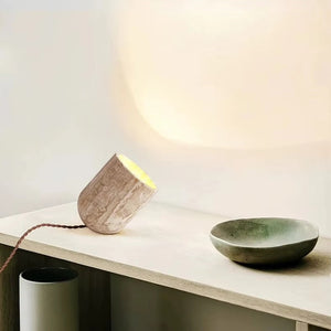 A sculptural Natural Stone Lamp, crafted from yellow cave stone in a small cylindrical shape, casts a warm glow on a wooden surface beside a round greenish-grey ceramic dish. Its graceful cord extends from the base.