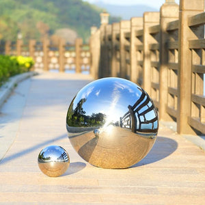 Two Stainless Steel Gazing Balls of different sizes are displayed on a paved walkway beside a wooden railing, with trees and mountains in the background.