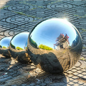 Three Stainless Steel Gazing Balls rest on a patterned stone surface, reflecting the sky and a nearby building with red accents.