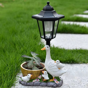 Solar Goose Lamp—a black outdoor lamp post adorned with a goose and goslings figurine, featuring a small potted plant and lush green grass in the background. This enchanting piece of garden decor is perfect for adding charm to your cottage garden.