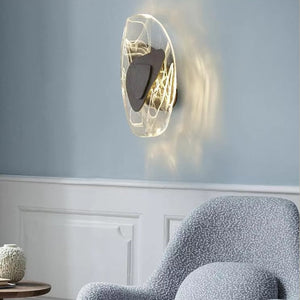 The Luxury Crystal Wall Lamp, featuring a sculptural glass and metal design, casts light above a curved, patterned armchair set against a pale blue wall.