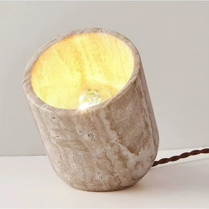 The Natural Stone Lamp, crafted from yellow cave stone, emits a warm light as it rests at an angle on a white surface, with a twisted brown power cord extending from the back.
