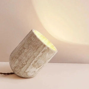 The Natural Stone Lamp, sculpted from yellow cave stone, leans at an angle on a flat surface and casts a warm, diffused light onto the beige wall behind it.