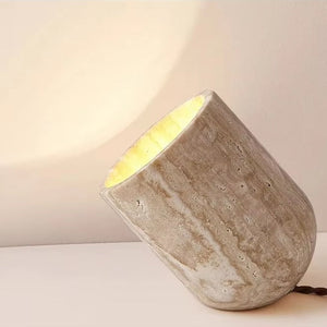The Natural Stone Lamp features a cylindrical, sculptural table lamp design inspired by the organic beauty of yellow cave stone. Tilted on its side, it casts a warm light onto beige walls and surfaces, seamlessly blending industrial form with natural inspiration.