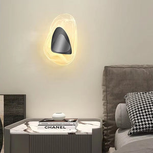 A Luxury Crystal Wall Lamp with an abstract design is mounted above a gray upholstered bed, positioned next to a marble nightstand decorated with a book and a houndstooth-patterned pillow.
