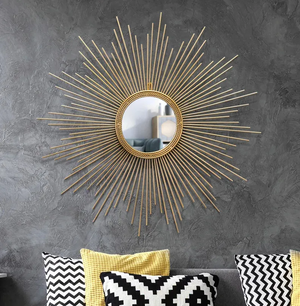 The Luxe Sunray Wall Mirror with a gold sunburst frame is mounted on a textured gray wall above a sofa with black, white, and yellow cushions, serving as a striking focal point in the room’s interior design.