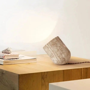 A sculptural Natural Stone Lamp, crafted in a cylindrical wooden form, lies at an angle on a wooden table beside a stack of books and a seashell.