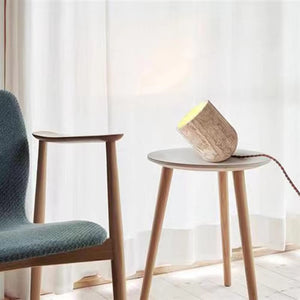 A wooden chair sits next to a small round table topped with the Natural Stone Lamp, all set before sheer white curtains.