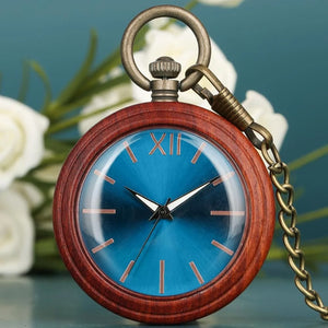 The Custom Vintage Antique Quartz Pocket Watch features a handmade wooden case, blue face, gold hour markers, and a bronze chain, shown resting against a blurred floral background.