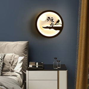 The Minimalist Wall Lamp, with its round design and bird-and-branch silhouette, emits a gentle and warm light that enhances the ambiance of a contemporary bedroom featuring a bed, cushions, and a nightstand decorated with bottles.