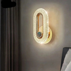 The Luxury Wall Sconce, an oval-shaped, wall-mounted LED light with an intricate design, emits a warm glow and is attached to a grey wall beside dark curtains, with a partial view of a bed.