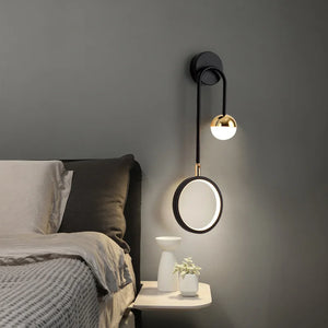 A stylish Hanging Circle Wall Light with a sleek black metal arm illuminates a bedside table adorned with two small vases and a plant, next to a bed featuring a gray headboard and neutral bedding. The modern design effortlessly incorporates geometric forms to elevate the space.