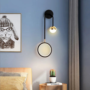The Hanging Circle Wall Light, a modern black sconce with two lights and a black metal arm, illuminates a room with blue walls, a framed animal print, and a bedside table holding a small potted cactus.
