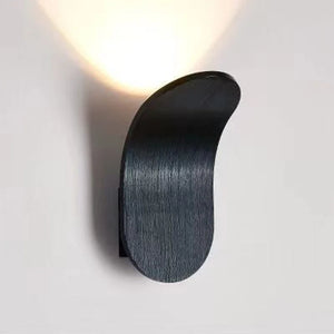 The Curved Wall Light, a Nordic-inspired wall lamp featuring a brushed metal design in black, emits light upwards and is mounted on a white wall. Perfect for living spaces seeking modern light fixtures that seamlessly blend elegance and functionality.