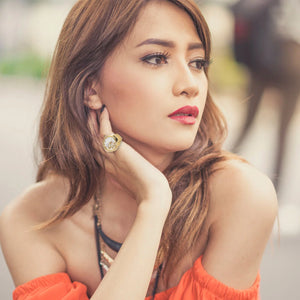 A woman with long brown hair and red lipstick rests her chin on her hand, wearing an off-shoulder orange top and the Butterfly Ring Watch, gazing thoughtfully to the side.