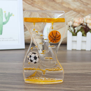 The Dual Color Liquid Hourglass, showcasing a black-and-white soccer ball and an orange basketball in a transparent liquid spiral, is beautifully displayed on a wooden surface with a small plant and framed picture in the background, creating a visually stunning experience.