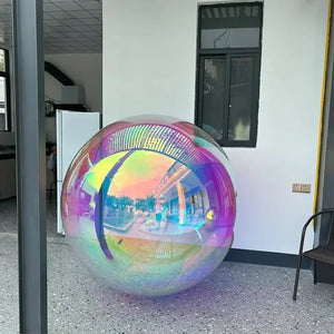 The Inflatable Gazing Ball, large, transparent, and iridescent, sits on a patio by a white building with windows and a chair, bringing unique garden decor charm to the space.