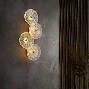 Four Amber Deco Wall Sconces with circular glass discs emit a warm glow on a gray textured wall beside dark pleated curtains.