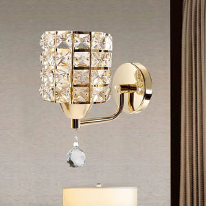 The Luxury Cylinder Wall Light is a gold crystal wall light with a grid-patterned shade and single hanging crystal, mounted on a beige wall next to a curtain.