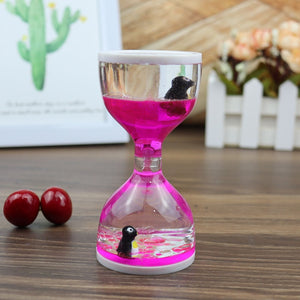 The visually stunning Dual Color Liquid Hourglass features a striking display of pink liquid and tiny black figures encased within it, all set on a wooden surface. Beside the hourglass are small décor items and scattered red beads, while a framed cactus image is softly blurred in the background.
