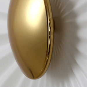 A close-up of the Origami Circle Wall Light, a smooth, oval-shaped gold accent, centered on a white ribbed surface with radial patterns.