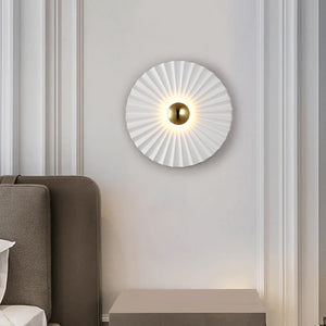 The Origami Circle Wall Light, featuring a modern pleated white shade and central gold accent, is mounted above a bedside table beside a bed.