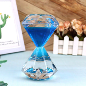 A Dual Color Liquid Hourglass, with its visually stunning diamond-shaped design, rests on a light blue tablecloth. It is surrounded by a wooden photo frame, a green plant, and a white flower pot filled with pink flowers.