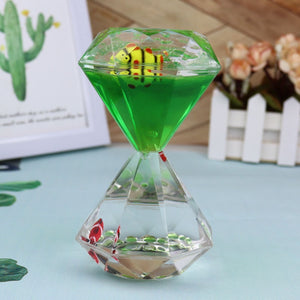 The Dual Color Liquid Hourglass features a green liquid-filled, hourglass-shaped container with a dual-color liquid display showcasing a decorative yellow and red striped object inside. The background includes a framed image of a cactus and some flowers in a white picket fence pot, creating a visually stunning experience.
