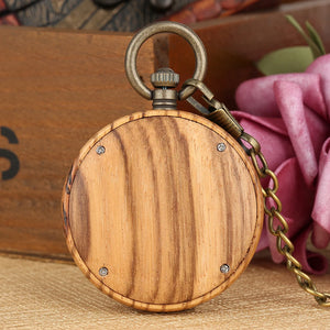 The Wooden Pocket Watch, featuring a closed wooden case and metal chain, is displayed on a textured surface beside pink artificial flowers and a wooden box.
