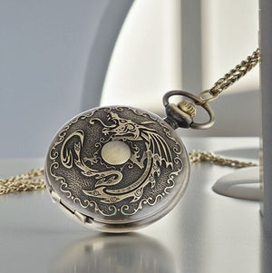 The Dragon Pocket Watch features a vintage design with a gold dragon and moon on the cover, comes with a matching gold chain, and is shown resting on a light-colored surface.