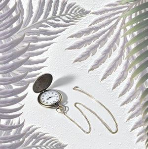 The Dragon Pocket Watch with a gold chain lies open on a textured white surface, surrounded by light purple and green fern leaves.