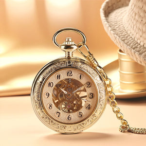 The Premium Golden Pocket Watch, featuring a skeleton dial and ornate details, is attached to a golden chain and rests on a beige surface near a woven hat.