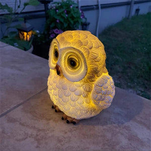 A Solar Owl Light, shaped like a yellow owl, glows on an outdoor table in the evening, serving as a charming garden decoration with plants and a lantern visible in the background.