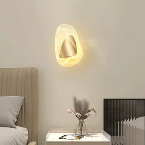 The Luxury Crystal Wall Lamp emits a warm glow above a nightstand adorned with a vase of dried plants, a magazine, and a decorative panel, positioned beside a beige upholstered bed.