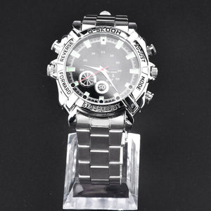 The Watch SpyCam is a silver metal wristwatch with a round black dial and buttons labeled "reverse," "forward," "adjust," and "mode." It discreetly records 1080P video and audio, shown on a clear stand against a black background.