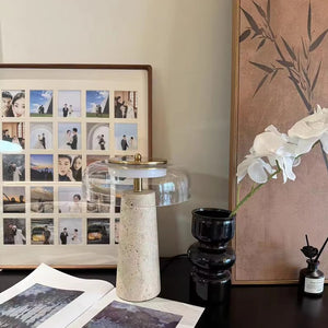 A desk featuring the Mushroom Desk Lamp, a framed photo collage, a black vase with white flowers, and an open magazine, positioned beside decorative brown artwork.