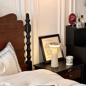 A Mushroom Desk Lamp, a floral mug, framed art, and various items sit on a bedside table next to a bed with white linens and a dark wooden headboard.