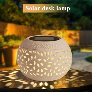 Round, white hollow iron lamp with a cutout leaf pattern, glowing on a table outdoors at dusk. Text reads “Solar Orb Lamp” at the top.