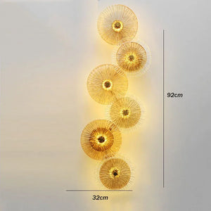 The Amber Deco Wall Sconce is a nature-inspired LED light with seven circular, fan-like elements arranged vertically. Measuring 92 cm tall by 32 cm wide, it casts a warm yellow glow.