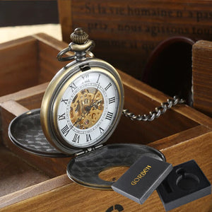 A Vintage Mechanical Pocket Watch with Roman numerals, open face, visible gears, and attached chain sits on a wooden box next to a black box marked "GOLDEN.