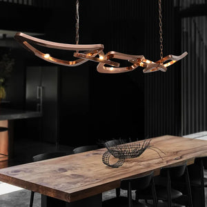 The Elegant Industrial Gallery Pendant, featuring sculptural curved rose gold metal, hangs above a large wooden dining table with black chairs in a dark, modern kitchen.
